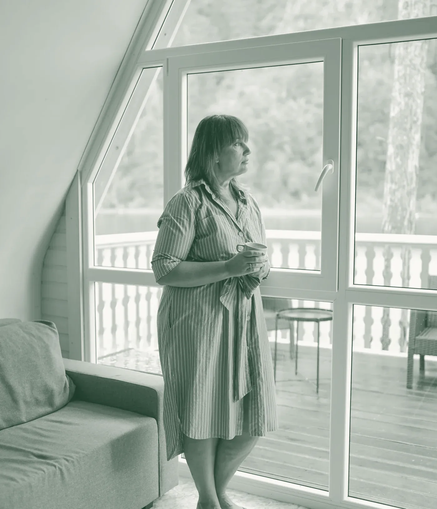 A woman in her forties stands by a bright window holding a cup of tea, looking out calmly